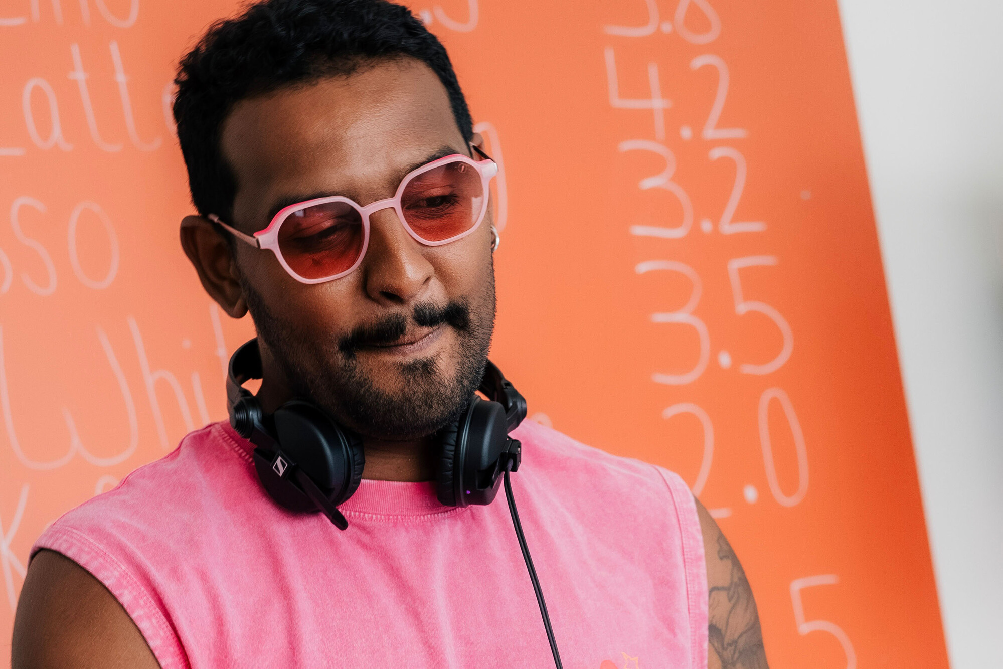 Kaffeebud steht mit pinkem Shirt und Kopfhörern um den Hals an den DJ-Reglern vor einer orangefarbenen Café-Tafel.