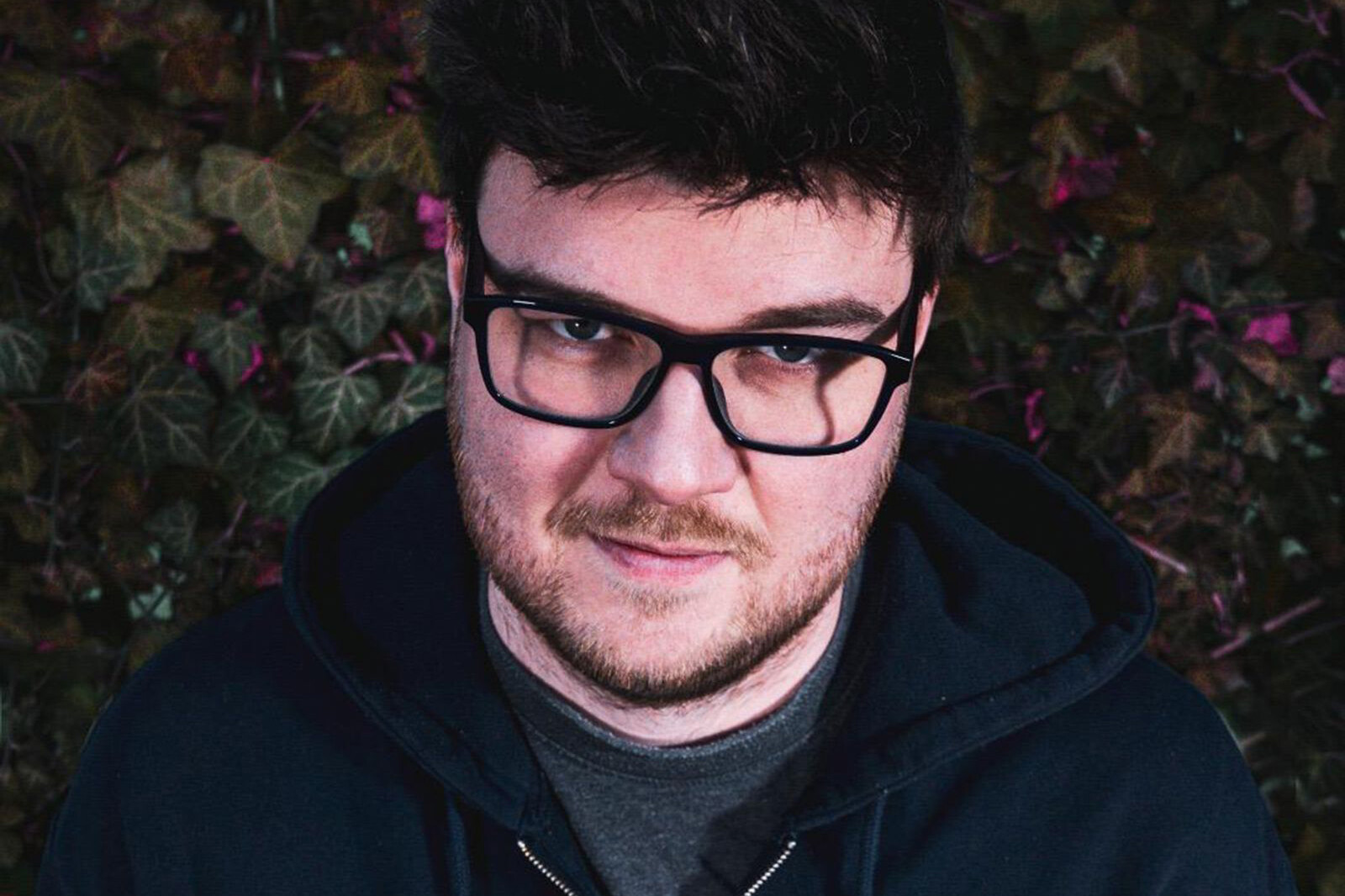 Puddlic blickt mit Brille und dunklem Kapuzenpulli vor einer Efeuwand direkt in die Kamera.